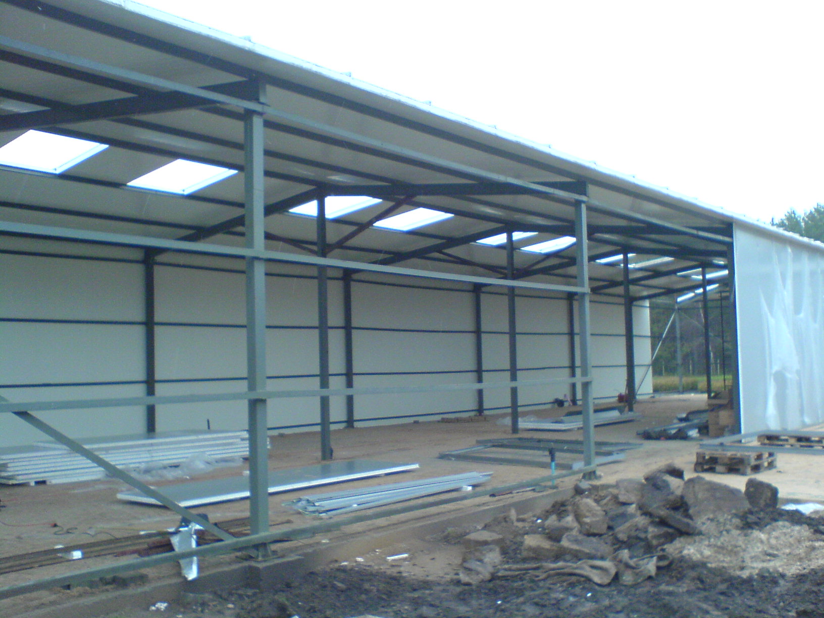 Wnętrze hali w trakcie budowy z widoczną konstrukcją stalową, panelami ściennymi i świetlikami w dachu, na podłożu porozrzucane materiały budowlane.